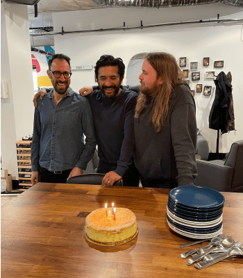 Jesse, Tris and Shea standing in front of their joint birthday cake with candles lit in the office.